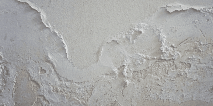 Close-up of peeling plaster and chipped white paint on interior wall, revealing rough textured surface