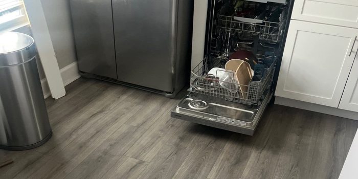Open dishwasher loaded with dishes next to a stainless steel refrigerator in a modern kitchen with gray wood floors.