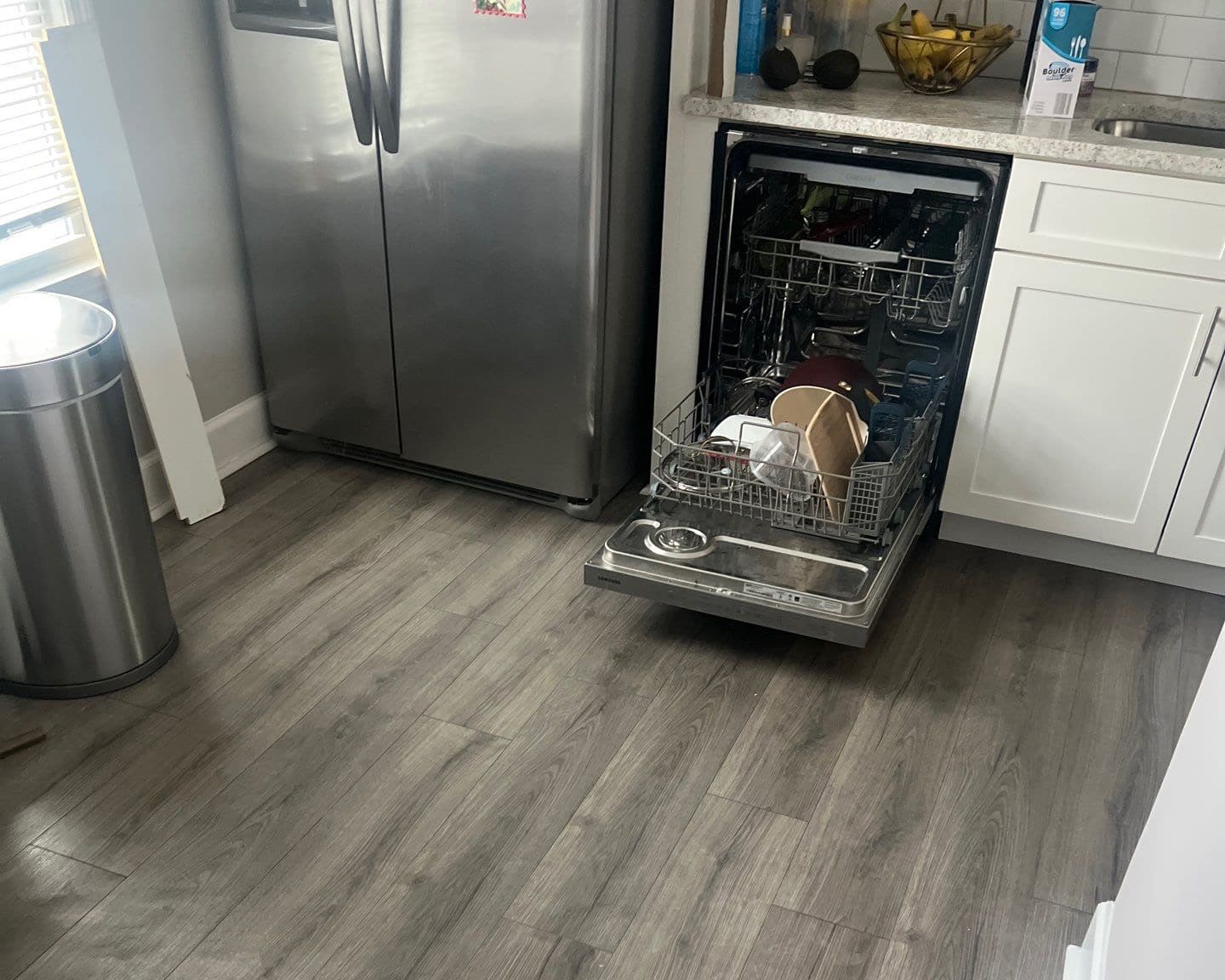 Open dishwasher loaded with dishes next to a stainless steel refrigerator in a modern kitchen with gray wood floors.