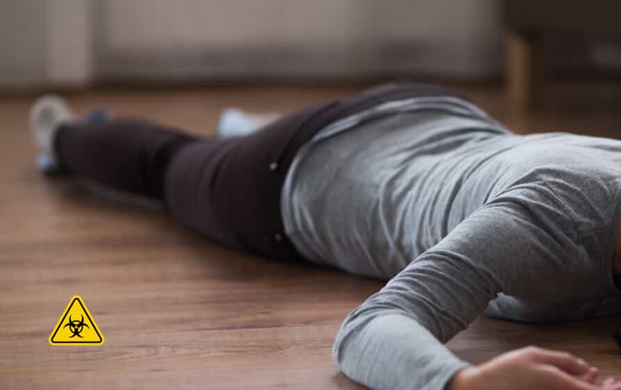 A person lying face down on a wooden floor indoors with a small biohazard warning symbol in the corner.