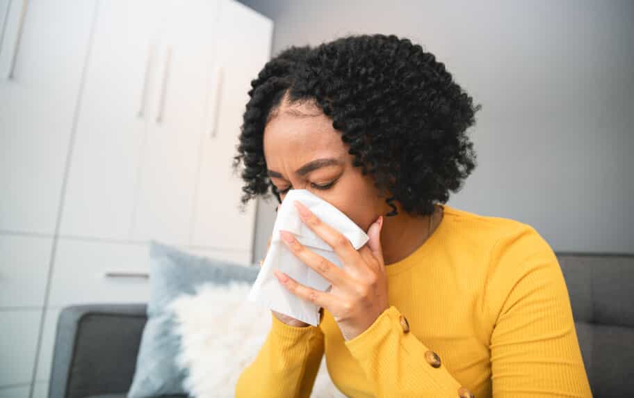A woman in a yellow sweater blowing her nose into a tissue on a couch in a living room.