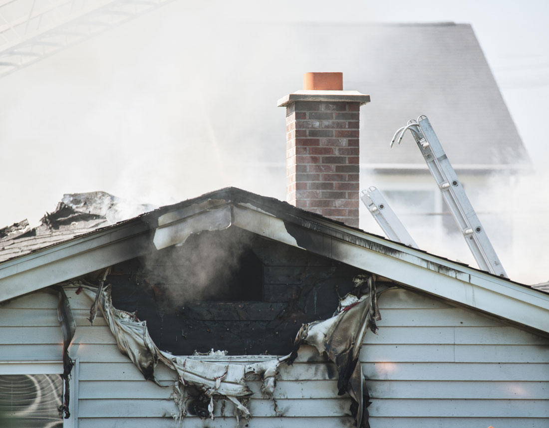 Smoke rising from fire-damaged gable and melted siding of a house with ladder by chimney