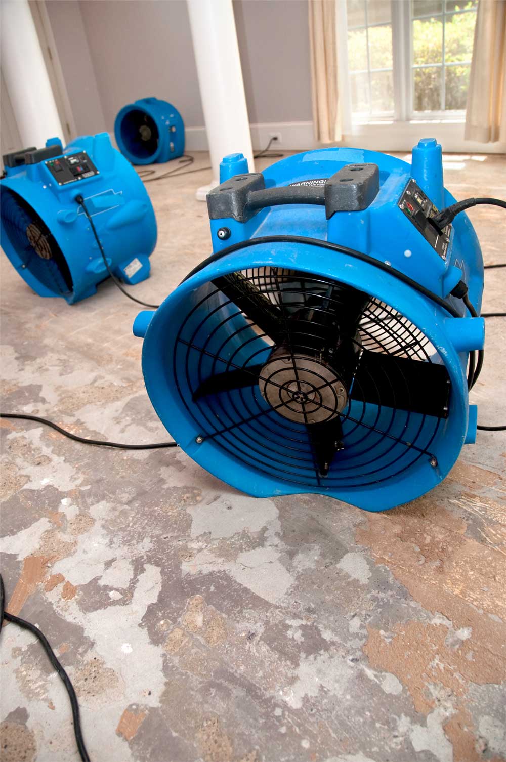 Three blue industrial air movers on a stripped concrete subfloor in a sunlit room drying during renovation