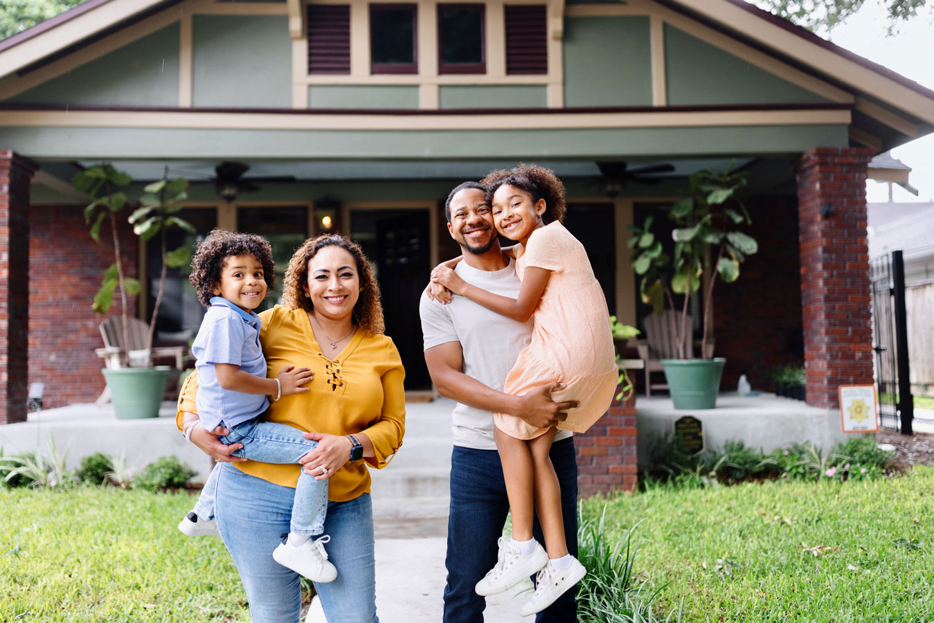 Smiling family of four—parents holding their two kids—stand on front lawn in front of their craftsman-style house