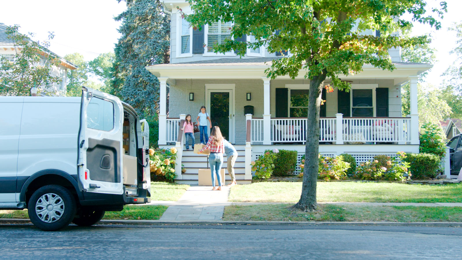 Family carrying moving boxes into a suburban house while a white moving van is parked on the street