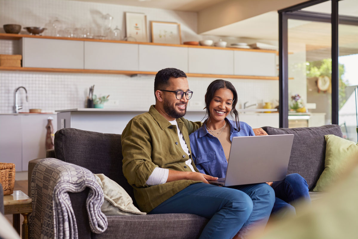 A man and a woman sitting on a couch, smiling while using a laptop in a bright modern living room/kitchen.