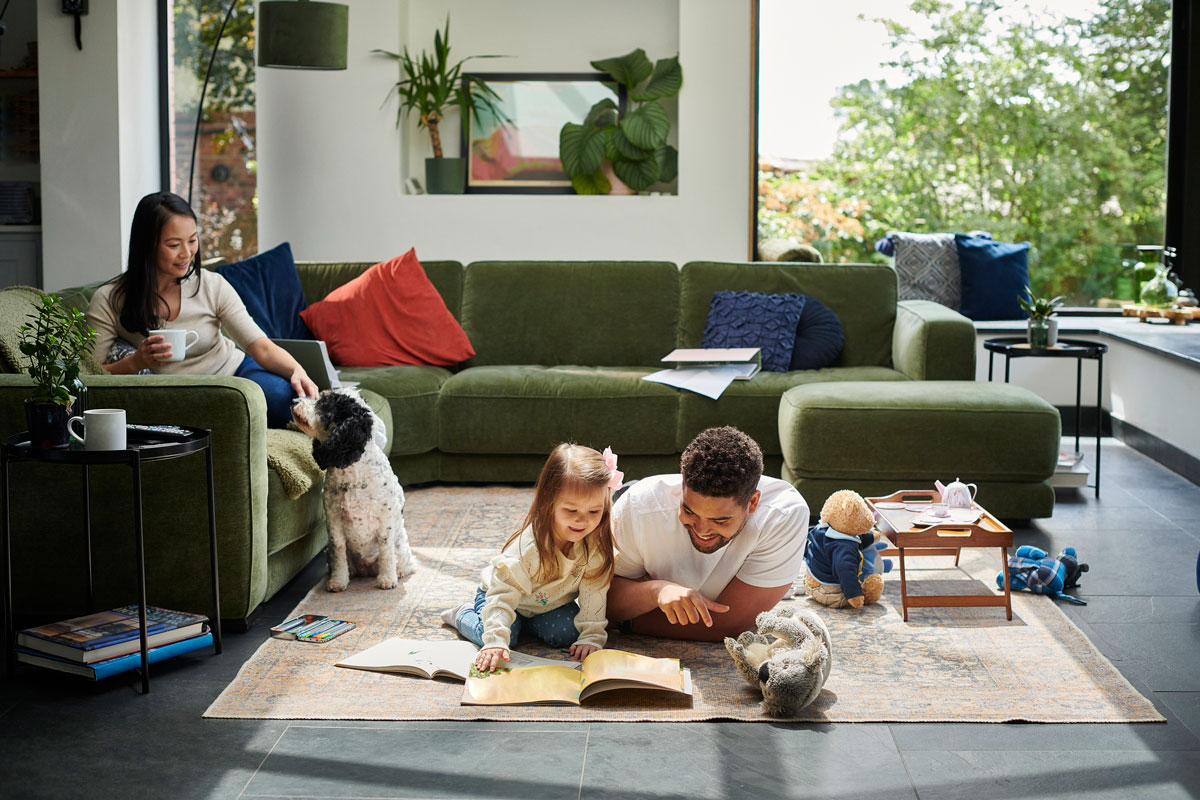 A man and young girl reading a picture book on the living room floor while a woman on the couch watches and a dog sits nearby