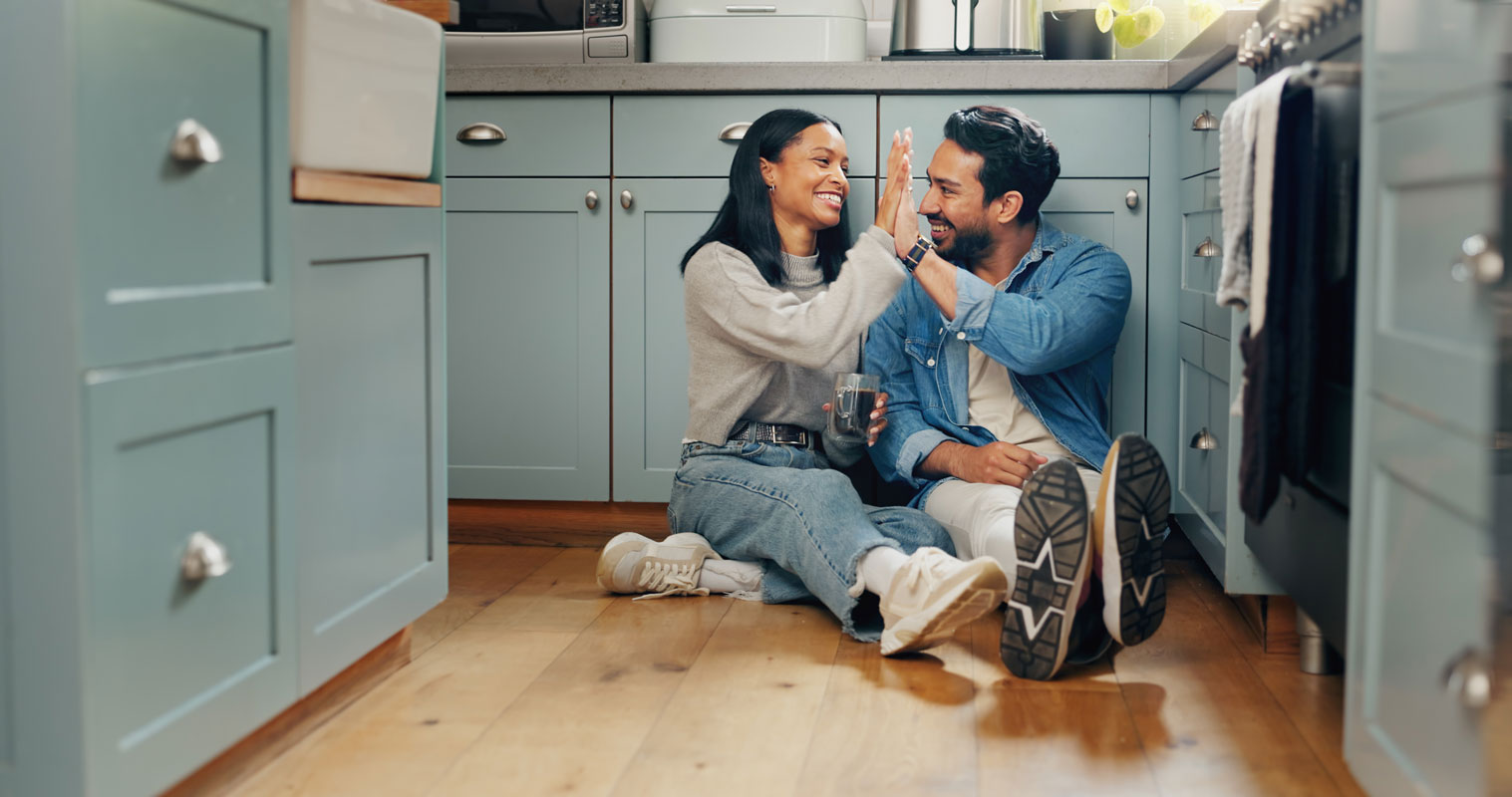 A smiling couple sitting on a hardwood kitchen floor, high-fiving while sharing a drink in a modern blue kitchen