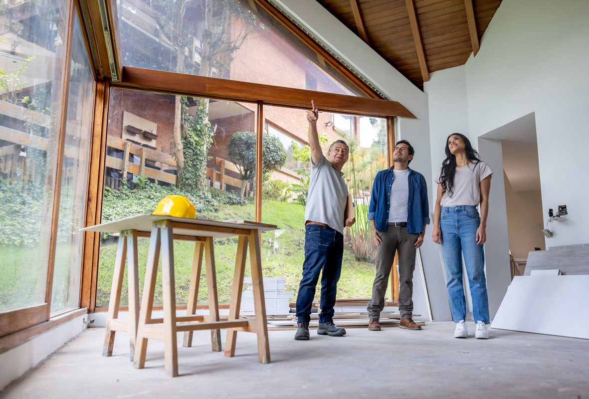 Contractor pointing at ceiling to a man and woman in sunlit living room; hardhat on table