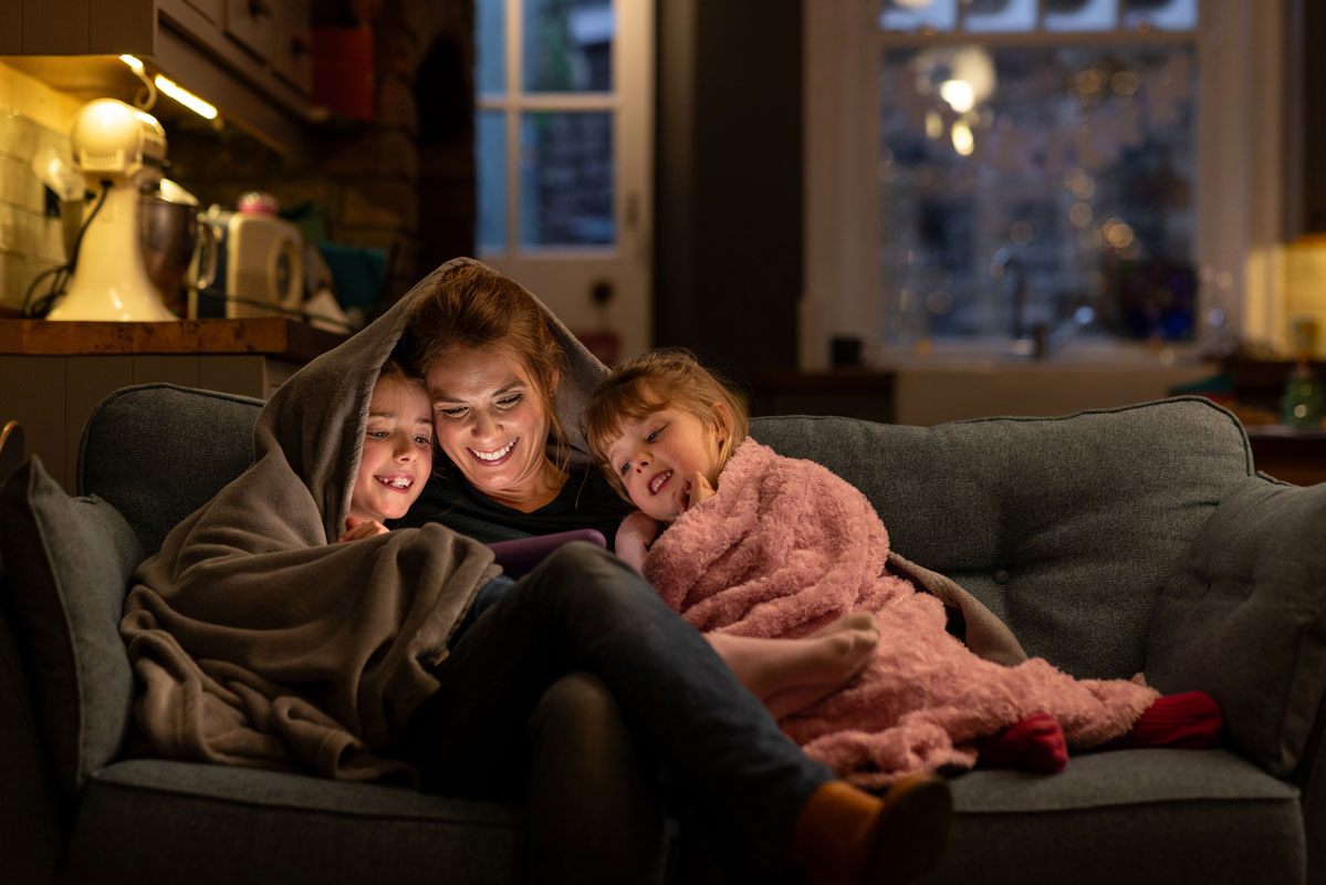 A woman and two children cuddled on a couch under blankets, smiling while looking at a tablet in a cozy living room.